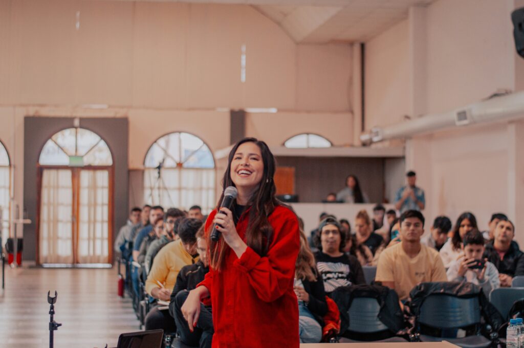 Dark haired woman in red with a microphone in front of a seated crowd