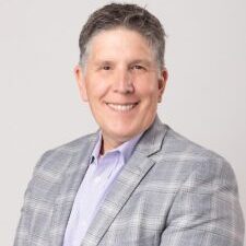 Smiling man with sandy hair wearing a lavender shirt and grey suit.